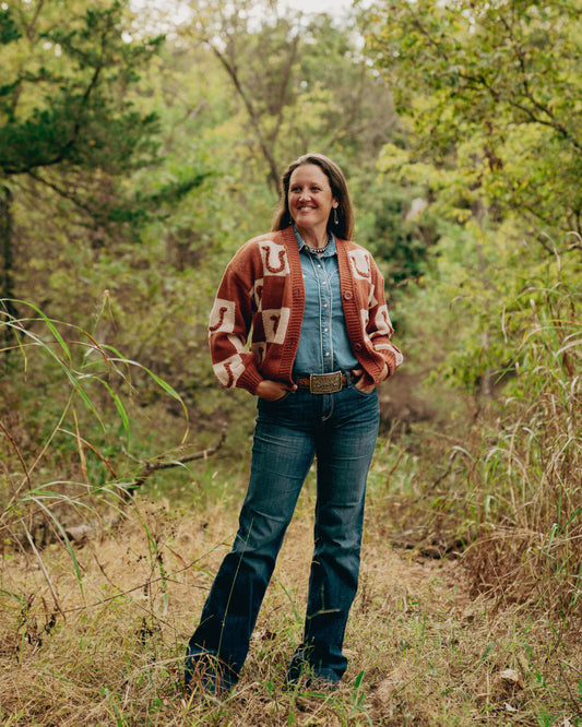 Woman standing in a forest wearing a patterned cardigan, denim jacket, and jeans.