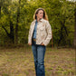 Woman standing in a forested area wearing a beige jacket and blue jeans.