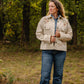 Woman standing in a forested area wearing a beige jacket and blue jeans.