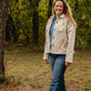 Woman standing in a forested area wearing a light-colored jacket with a floral design.