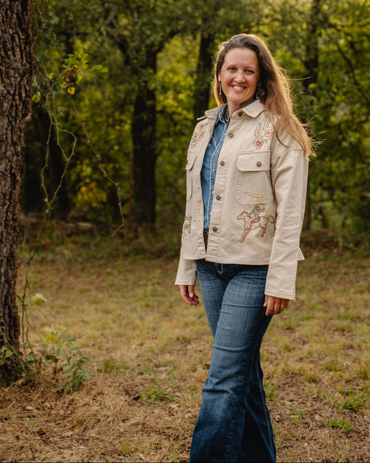 Woman standing in a forested area wearing a light-colored jacket with a floral design.
