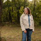 Woman standing in a forest wearing a beige jacket with floral patterns.