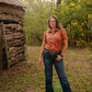 woman standing in woods wearing a short sleeved blouse and jeans with rustic cabin in the background