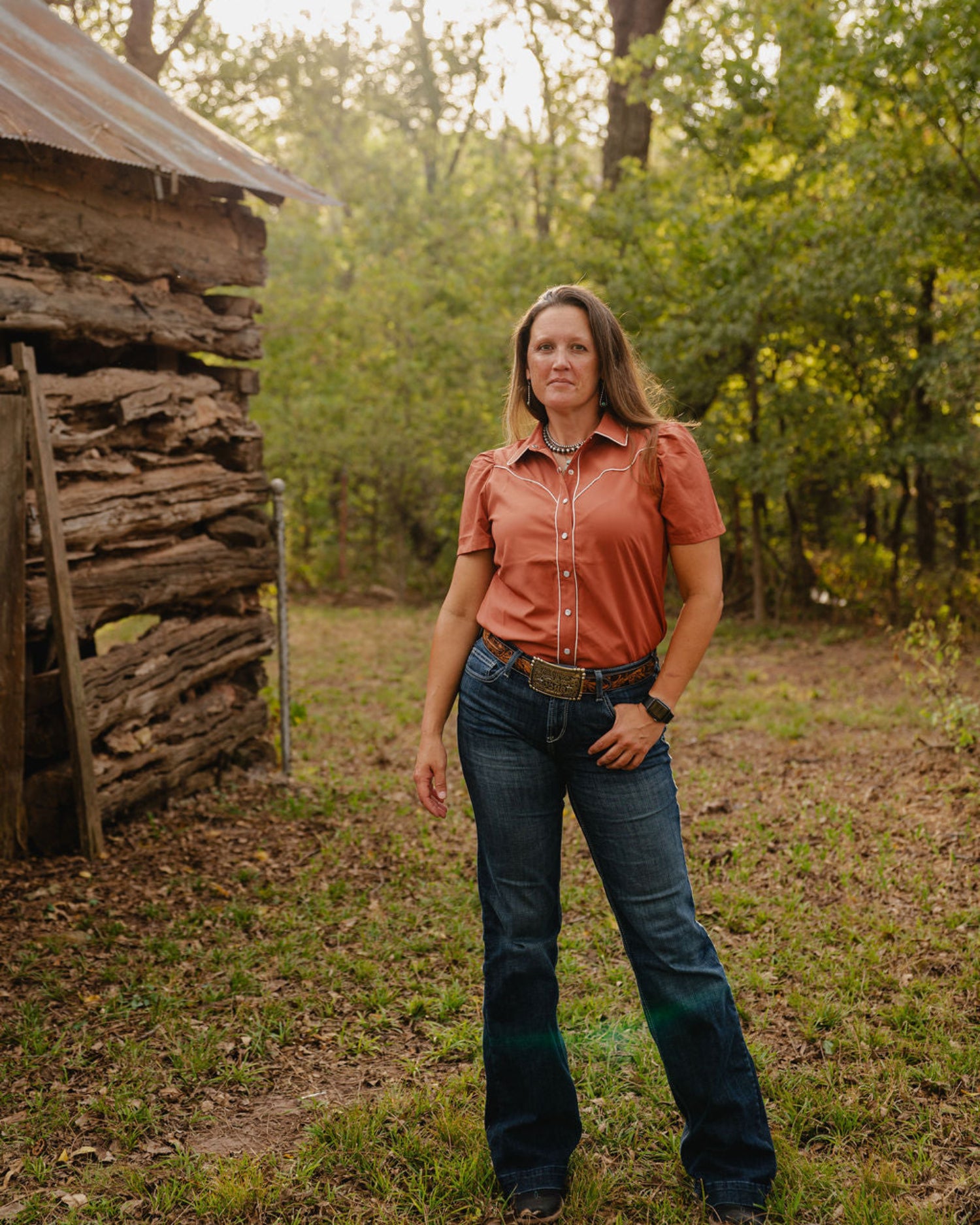 woman standing in woods wearing a short sleeved blouse and jeans with rustic cabin in the background