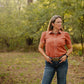 Woman standing in a forested area wearing a rust shirt and blue jeans.