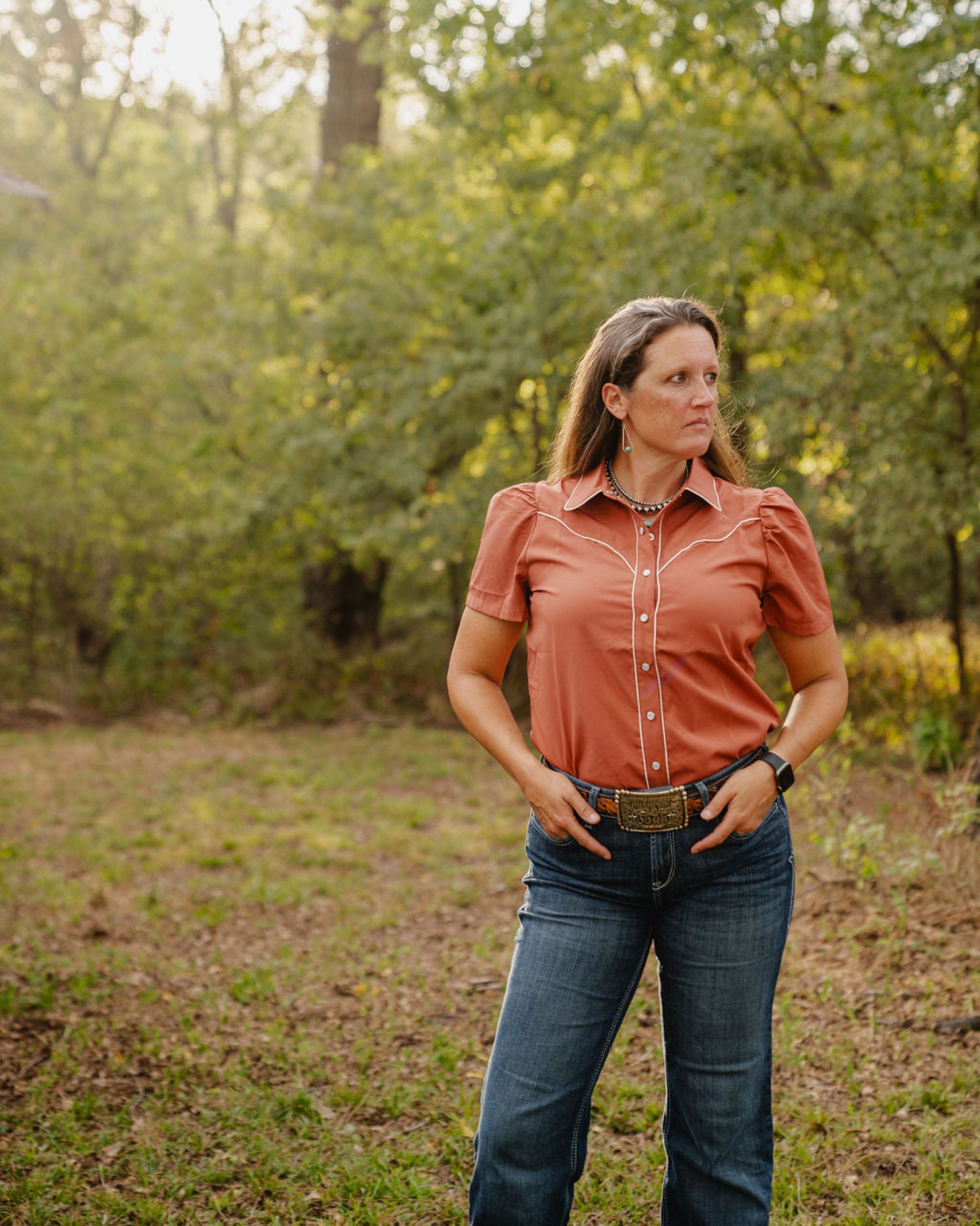 Woman standing in a forested area wearing a rust shirt and blue jeans.