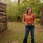 Woman in a rust-colored shirt and blue jeans standing in a natural setting with trees and a wooden structure.