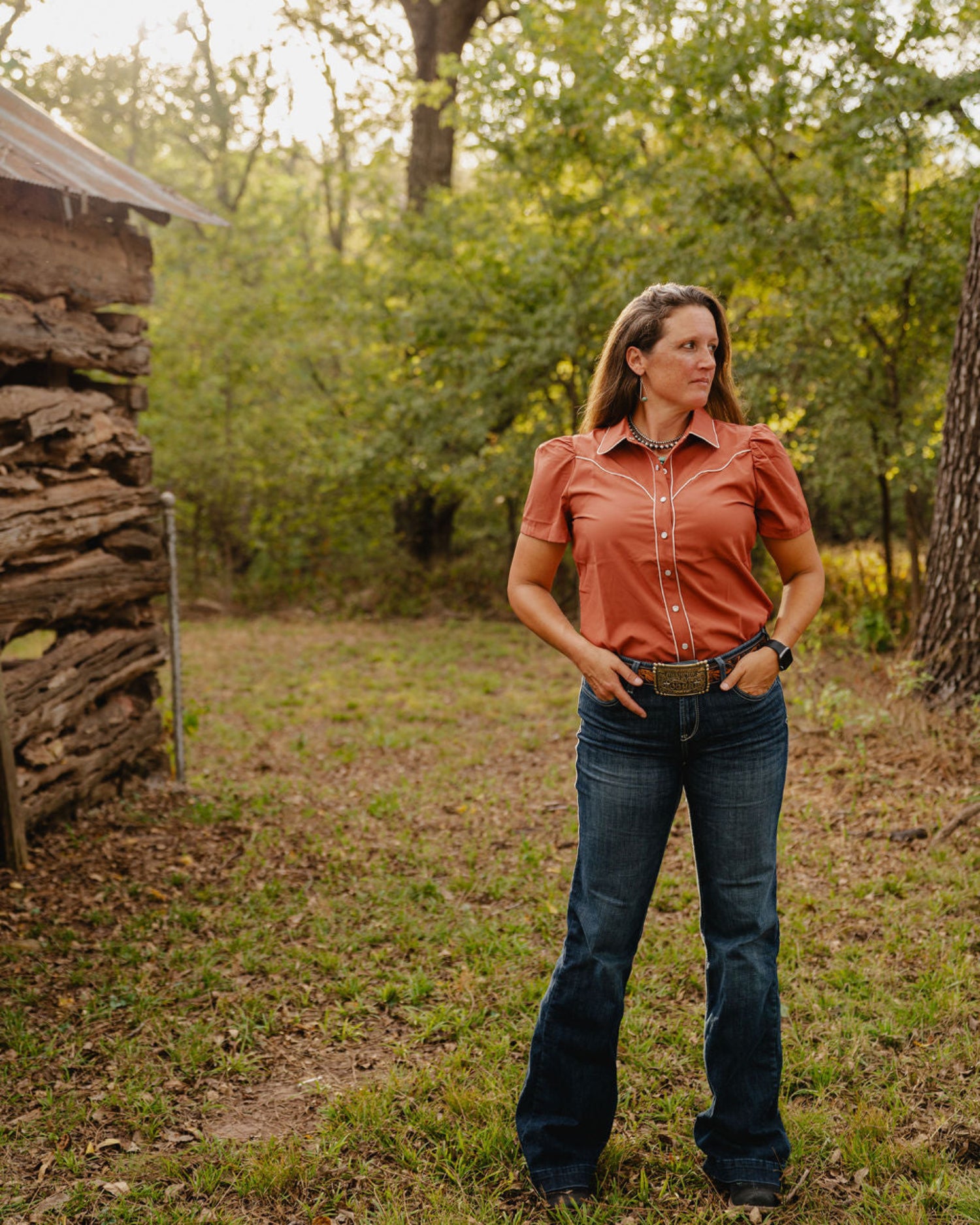 Woman in a rust-colored shirt and blue jeans standing in a natural setting with trees and a wooden structure.