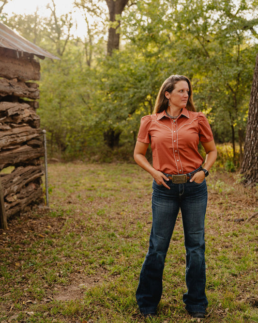 Woman in a rust-colored shirt and blue jeans standing in a natural setting with trees and a wooden structure.