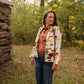 Woman standing outdoors in a natural setting with trees and a wooden structure.