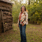 woman wearing jeans and a jacket standing in front of a rustic building with trees in background