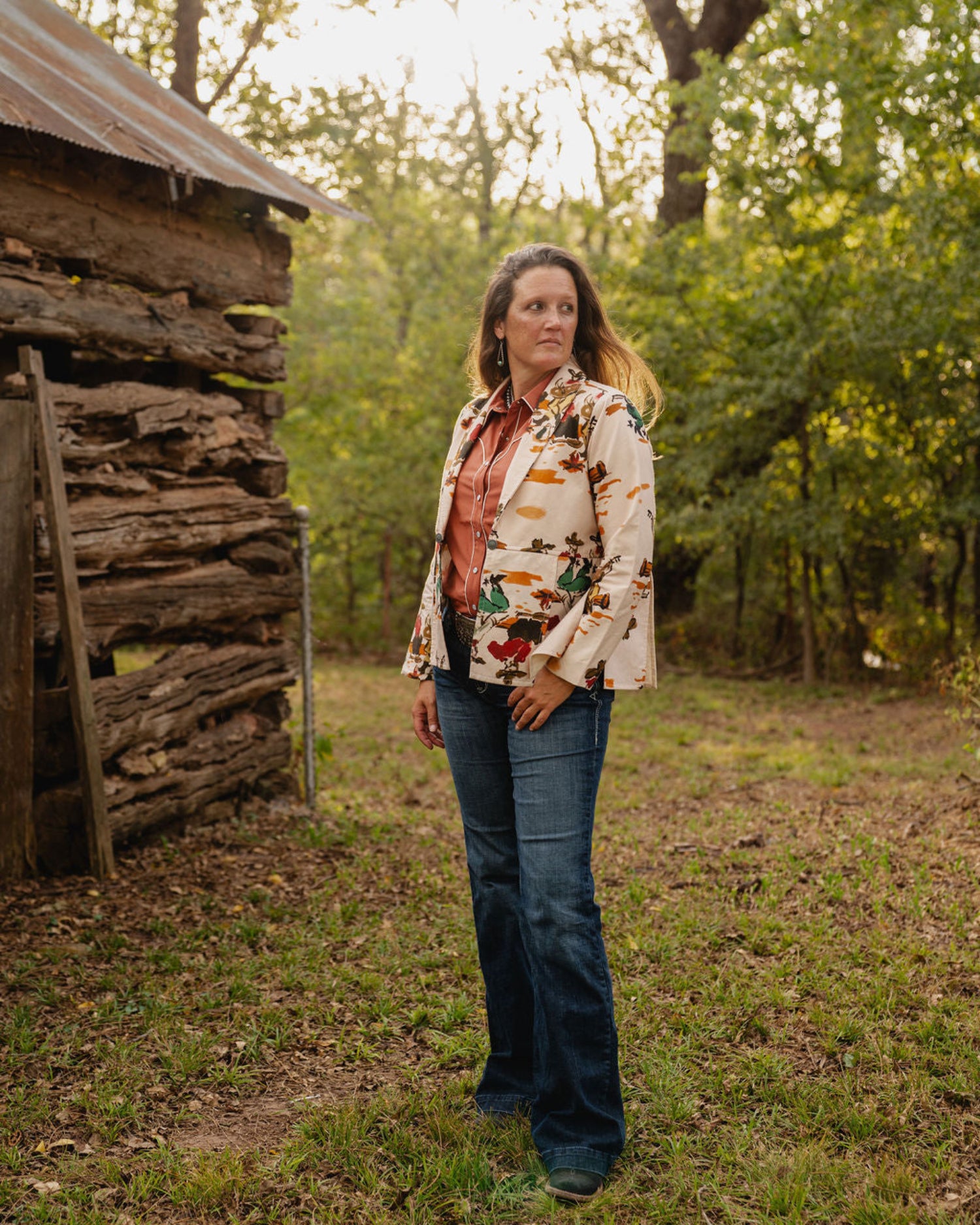 woman wearing jeans and a jacket standing in front of a rustic building with trees in background