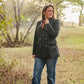 Woman wearing a dark jacket and jeans standing in a field with trees in the background