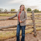 Woman standing by a wooden fence in an open field with a cloudy sky.