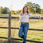 Woman standing by a wooden fence in a grassy field with trees in the background