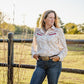 Woman standing in a field wearing a patterned blouse and jeans, with a wooden fence and trees in the background.