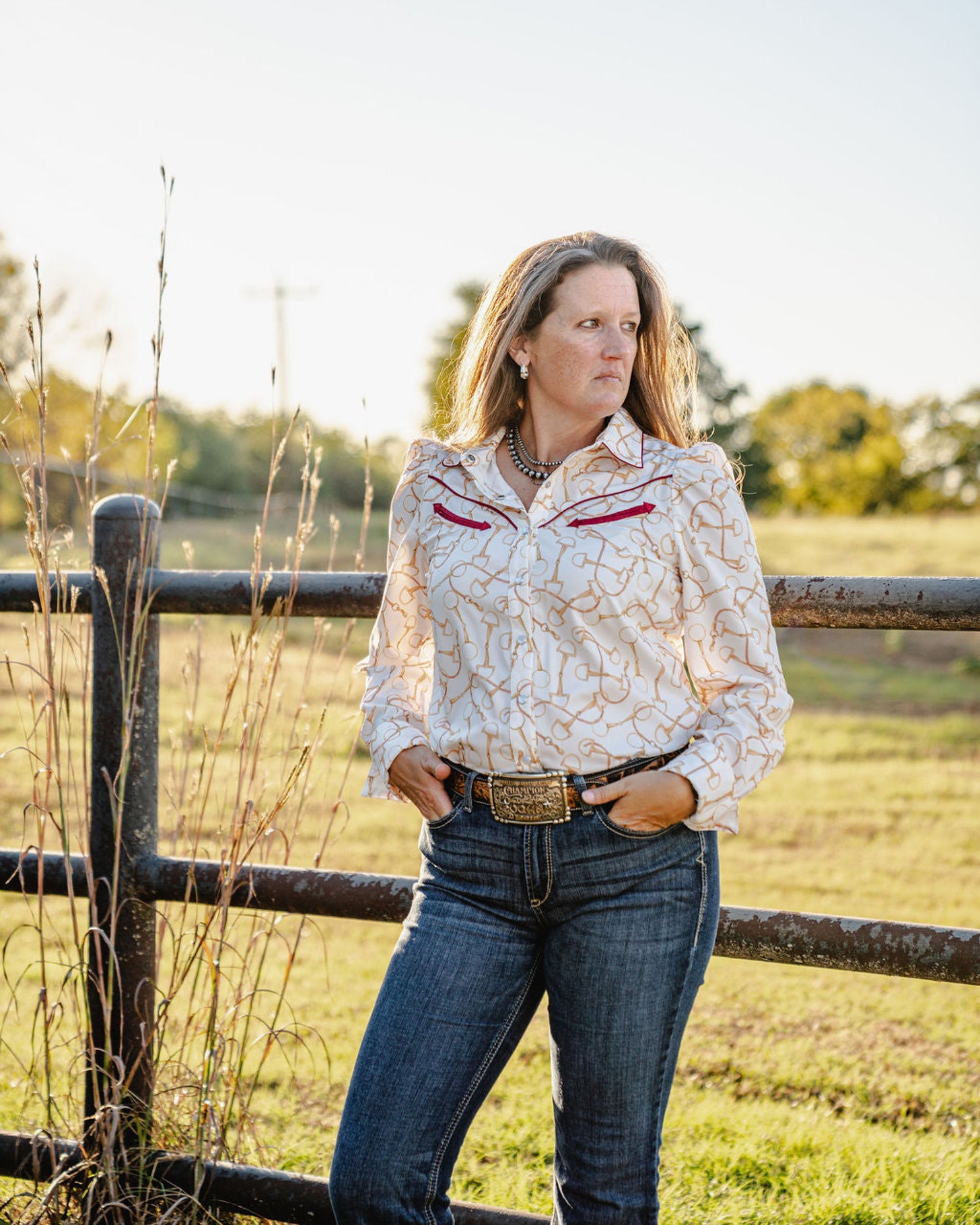 Woman standing in a field wearing a patterned blouse and jeans, with a wooden fence and trees in the background.