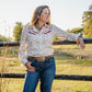 Woman in a patterned shirt and jeans leaning against a fence in a field.