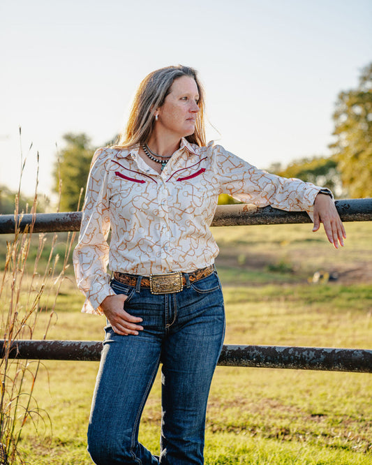 Woman in a patterned shirt and jeans leaning against a fence in a field.