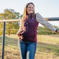 Woman standing in a field wearing a purple vest and jeans, with trees and clear sky in the background.