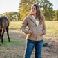 Woman standing in a field with a horse, wearing a beige jacket and blue jeans.