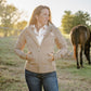 Woman standing in a field with a horse, wearing a beige jacket and blue jeans.