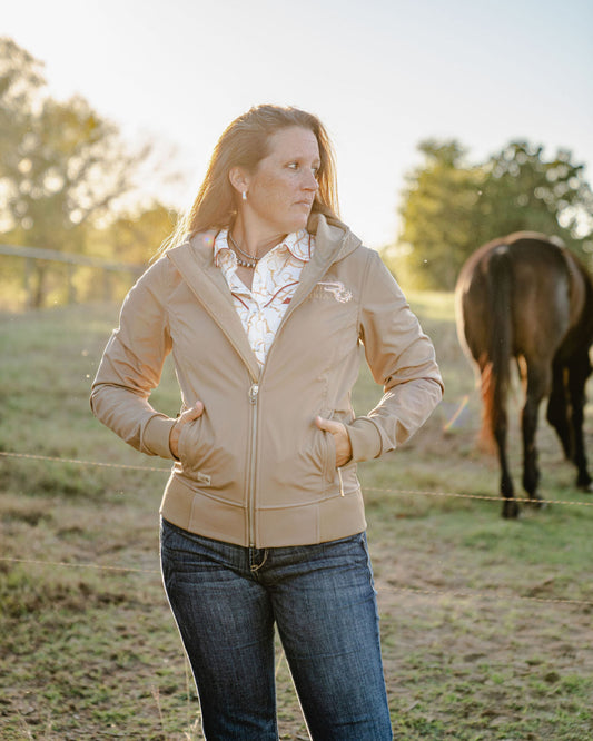 Woman standing in a field with a horse, wearing a beige jacket and blue jeans.
