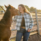 Woman standing next to a horse in an outdoor setting with a wooden fence and trees in the background.