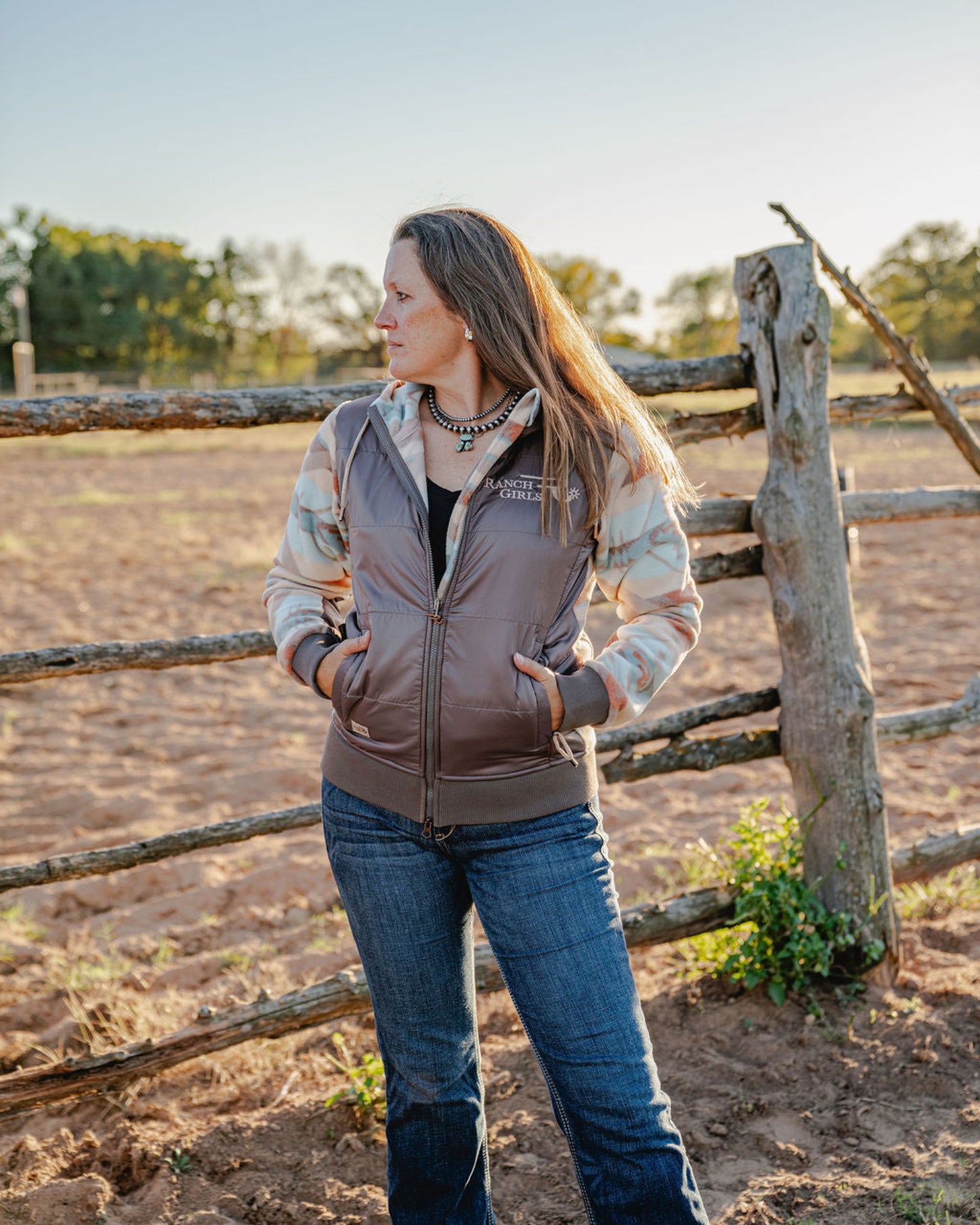 Woman standing in a field next to a wooden fence wearing a jacket with a logo.