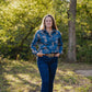 Woman wearing a blue floral shirt and jeans standing in a grassy area with trees.