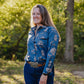 Woman wearing a blue floral shirt standing outdoors with greenery in the background