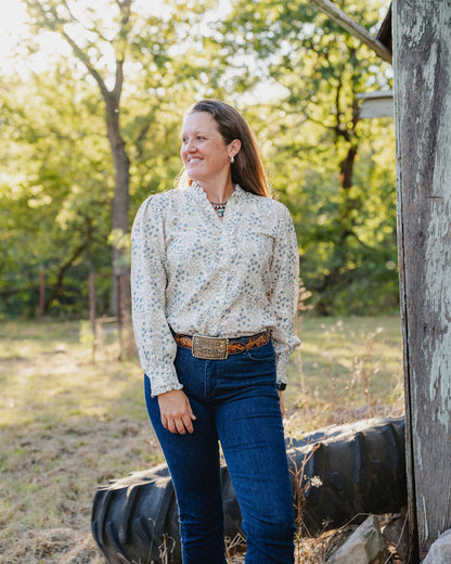 Woman standing outdoors in a natural setting with trees and a wooden structure.
