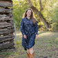 Woman in a blue dress standing in a natural setting with trees and a wooden structure.