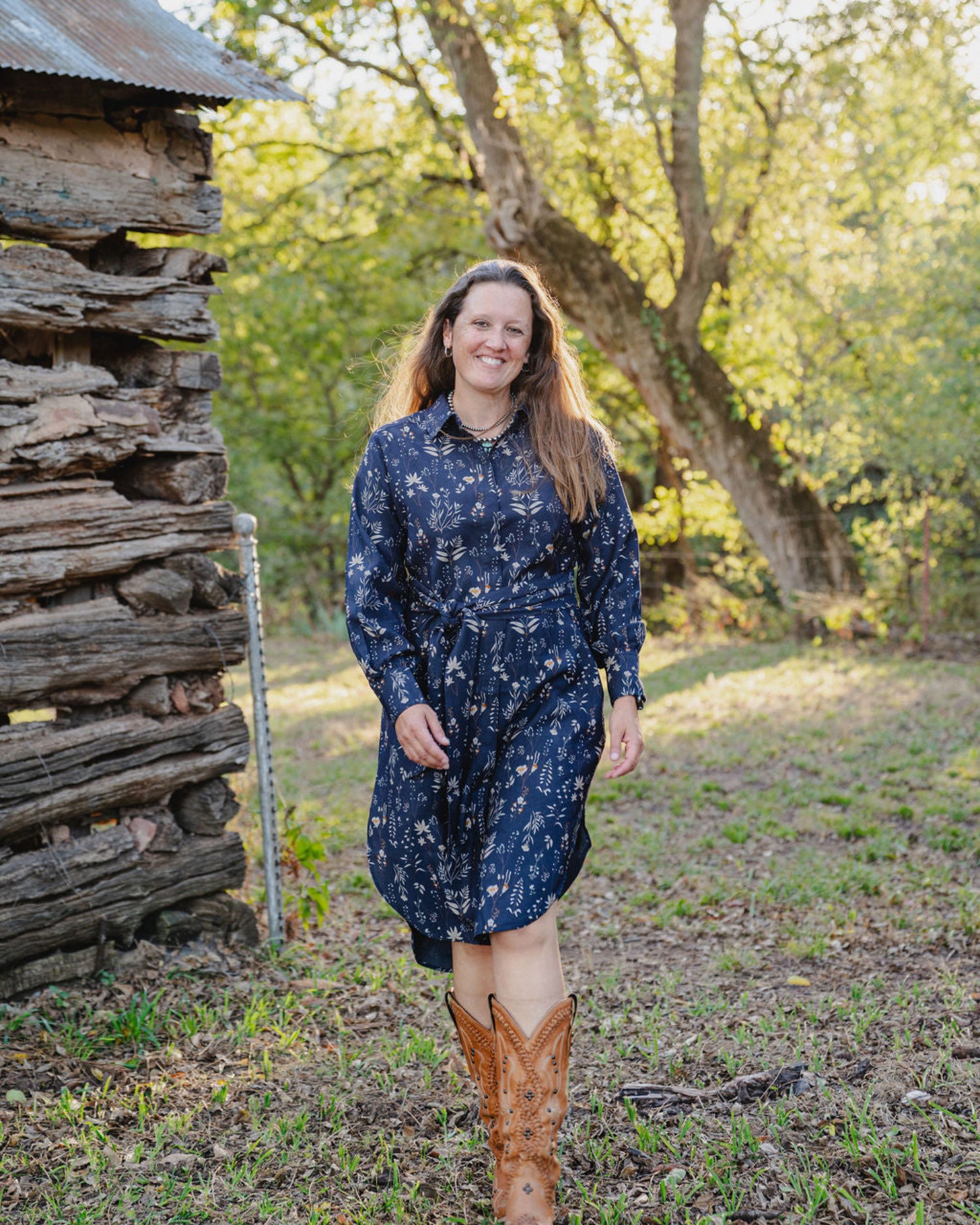 Woman in a blue dress standing in a natural setting with trees and a wooden structure.
