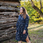 Woman in a blue floral dress standing in front of a rustic wooden cabin.