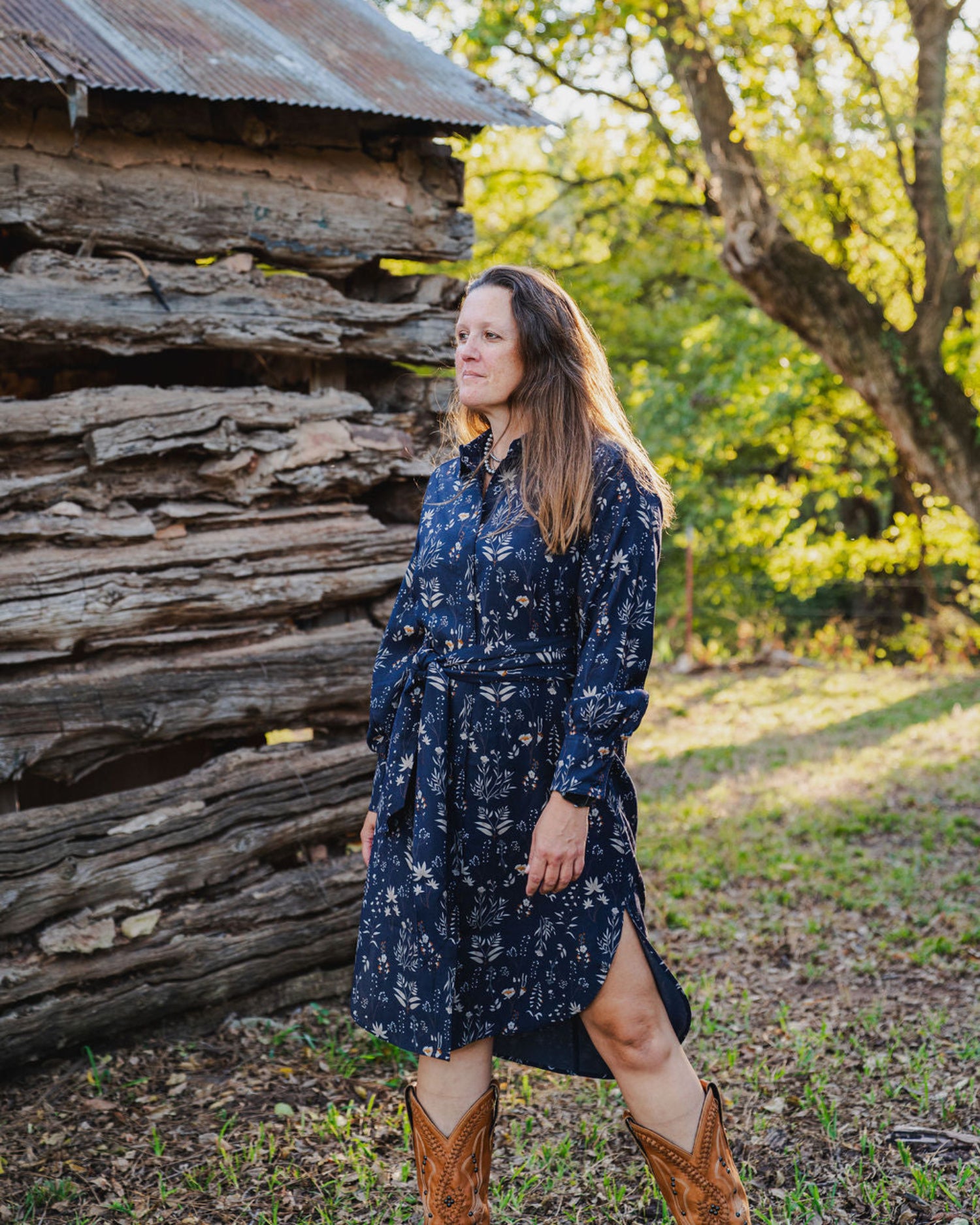Woman in a blue floral dress standing in front of a rustic wooden cabin.