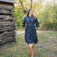 Woman in a blue floral dress and brown boots standing in a natural setting with trees and a wooden structure.