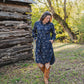 Woman in a blue floral dress and brown boots standing in front of a wooden cabin.