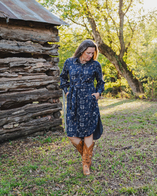 Woman in a blue floral dress and brown boots standing in front of a wooden cabin.