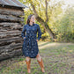 Woman in a blue floral dress and brown boots standing in a natural setting with trees and wooden logs.