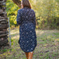 Woman in a navy floral dress standing in a forested area with a wooden structure.