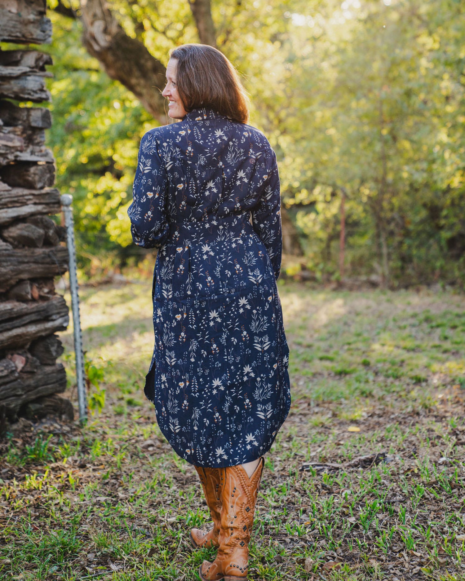 Woman in a navy floral dress standing in a forested area with a wooden structure.