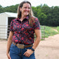 Woman wearing a patterned shirt and jeans standing outdoors with trees and a building in the background