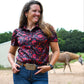 Woman in a patterned shirt and jeans standing in an outdoor setting with a horse in the background
