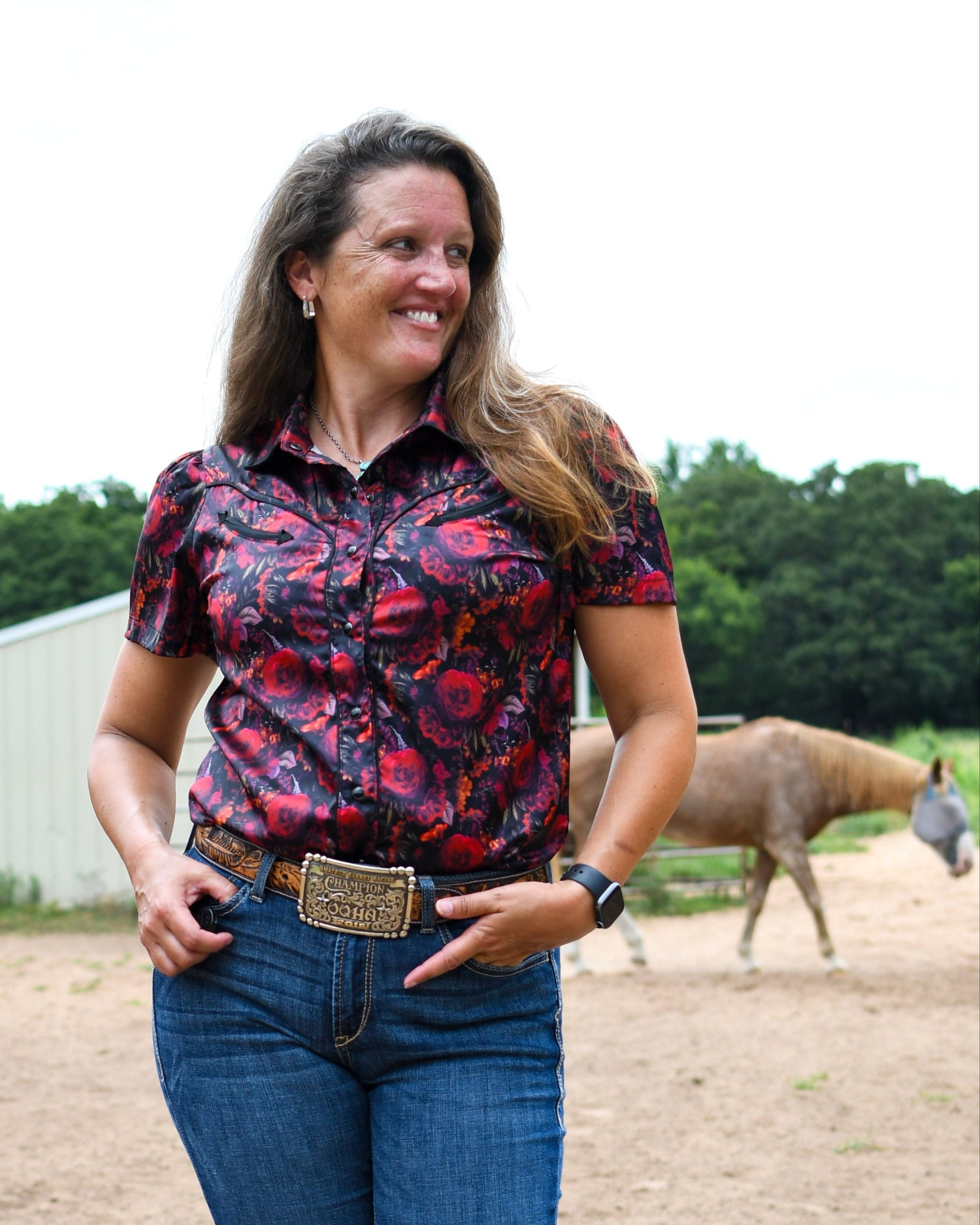 Woman in a patterned shirt and jeans standing in an outdoor setting with a horse in the background