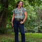 Woman standing outdoors in a forested area wearing a patterned shirt and jeans.