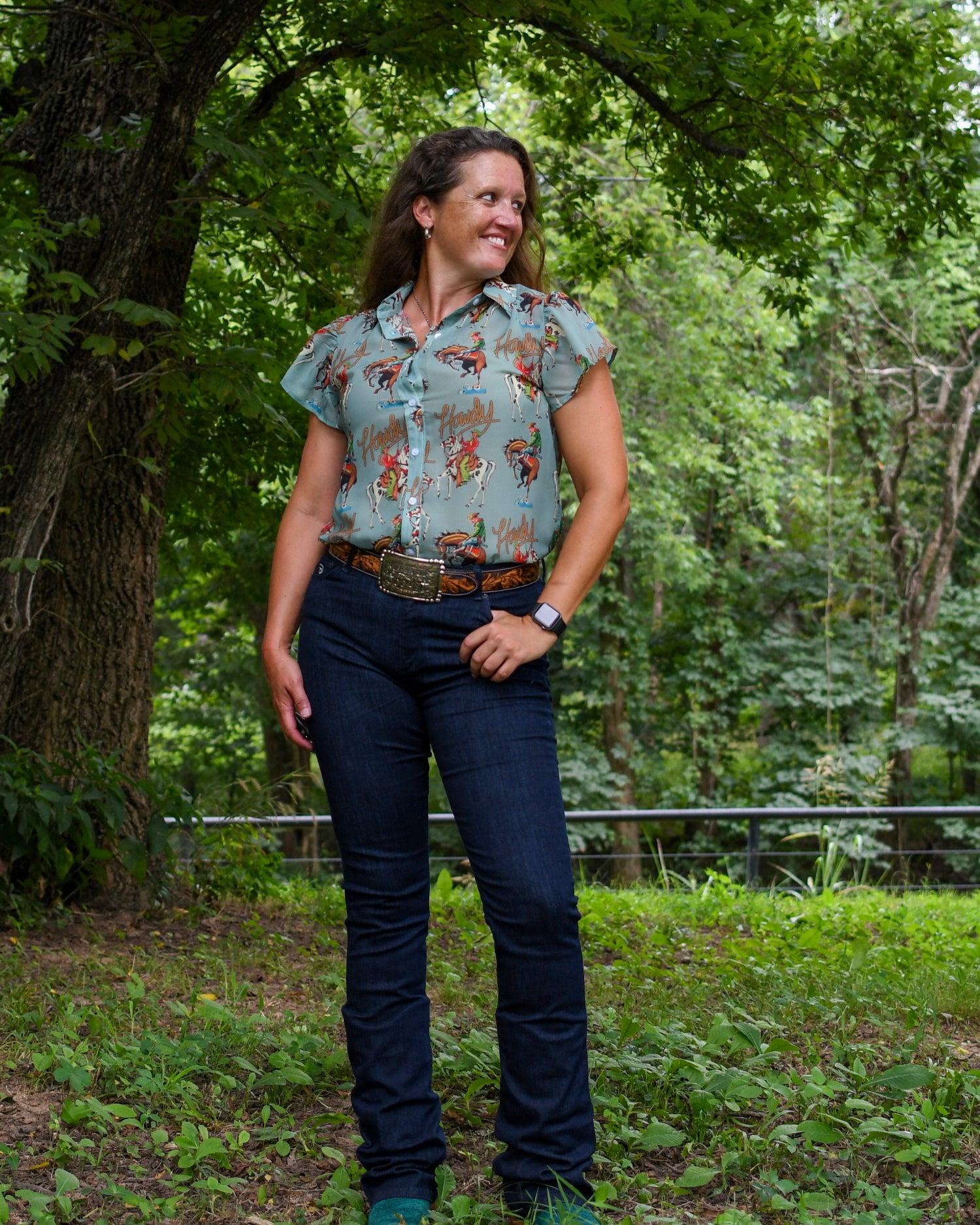 Woman standing outdoors in a forested area wearing a patterned shirt and jeans.