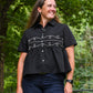 Woman wearing a black blouse with white embroidery in a forest setting