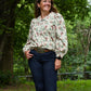 Woman standing outdoors in a natural setting wearing a floral blouse and dark jeans.