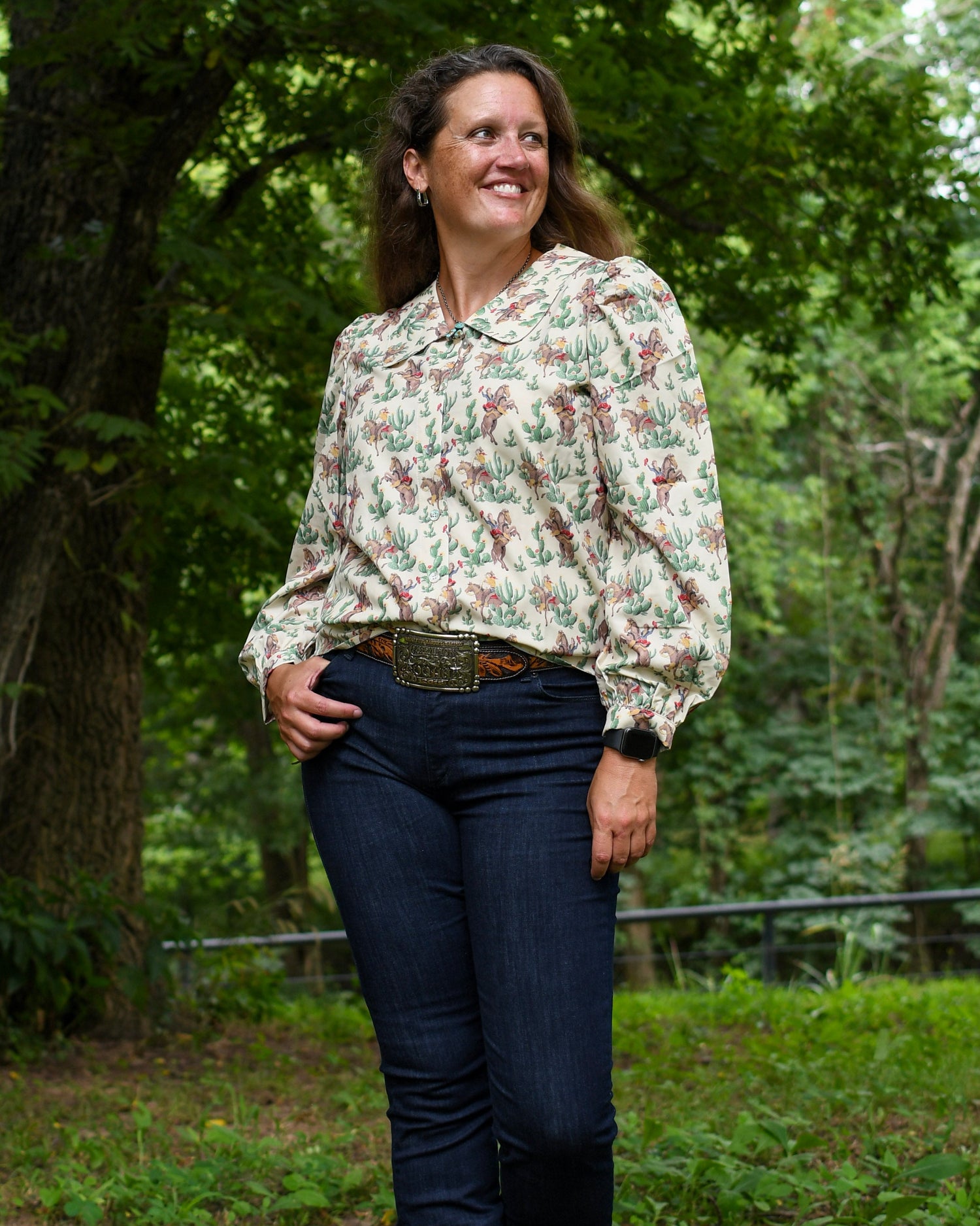 Woman standing outdoors in a natural setting wearing a floral blouse and dark jeans.